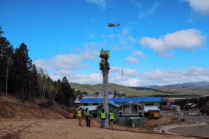 The crew readys the tower for its cross arm while the helicopter carries a tower section in the background.